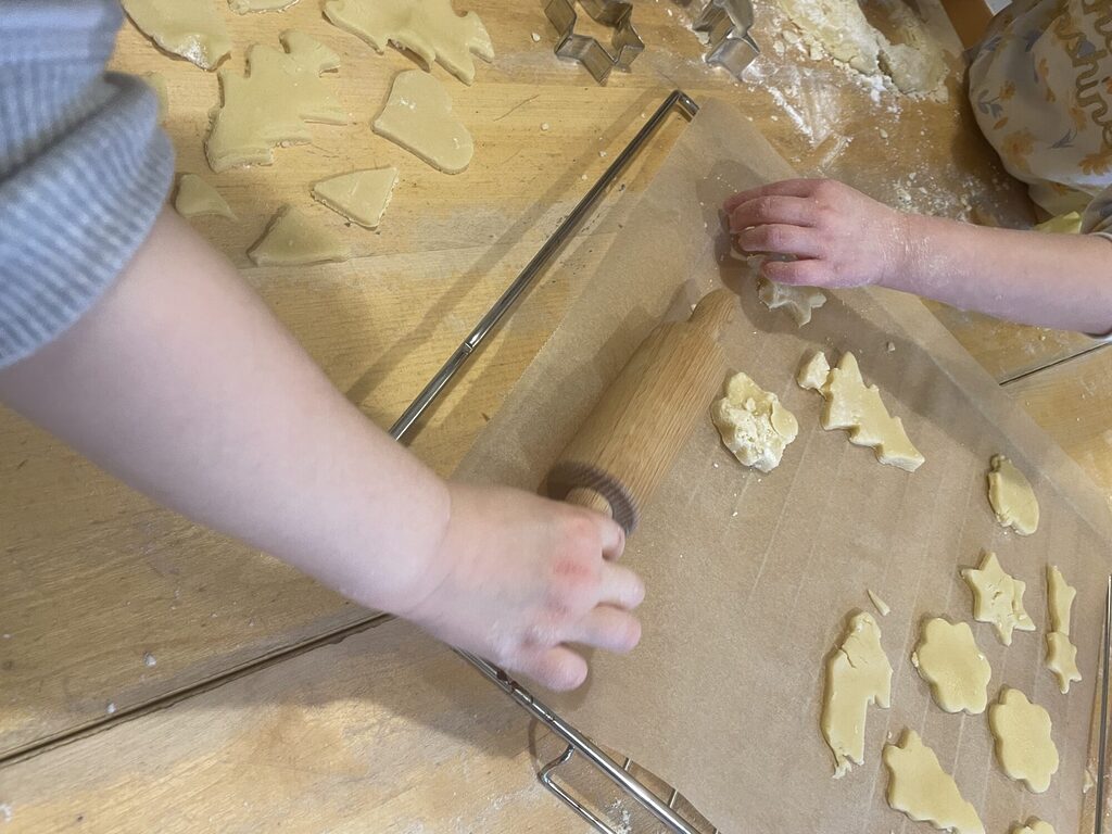 Kinderhände legen fertig ausgestochene Plätzchen auf ein Blech mit Backpapier. Eine andere Hand greift nach einem Nudelholz.