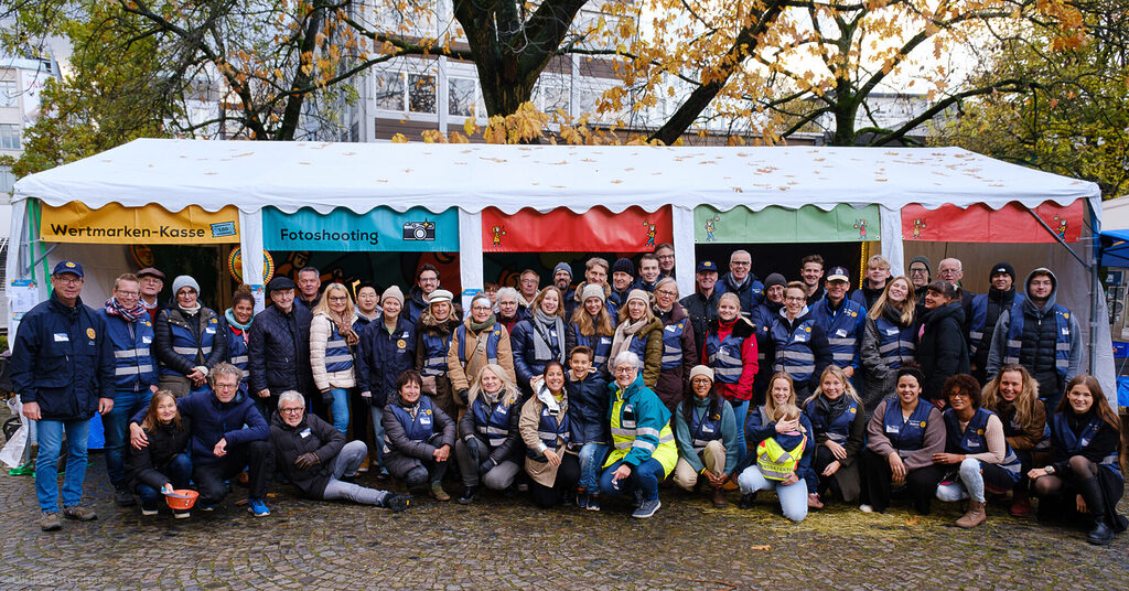 eine sehr große Gruppe an Ehrenämtern aus verschiedenen Clubs posiert für ein Gruppenfoto vor einem großen bunten Zelt des St.-Martins-Marktes. Alle tragen eine dunkelblaue Warnweste.