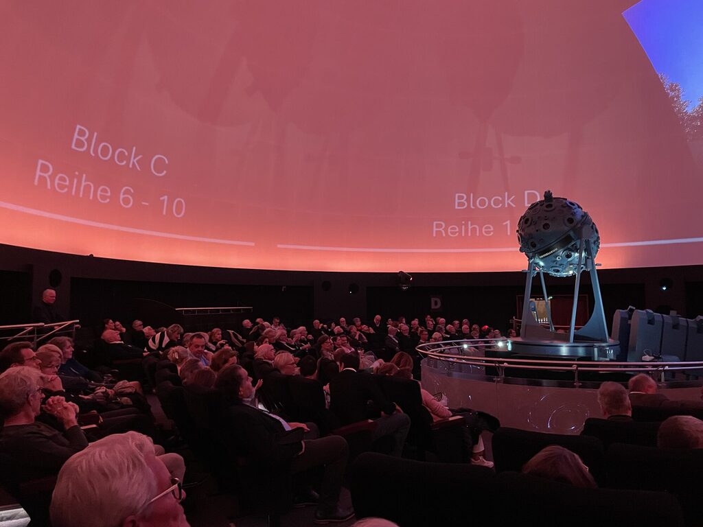Ein großes Publikum sitzt im Planetarium. Der große Projektor in der Mitte beleuchtet die runde Kuppel von innen.
