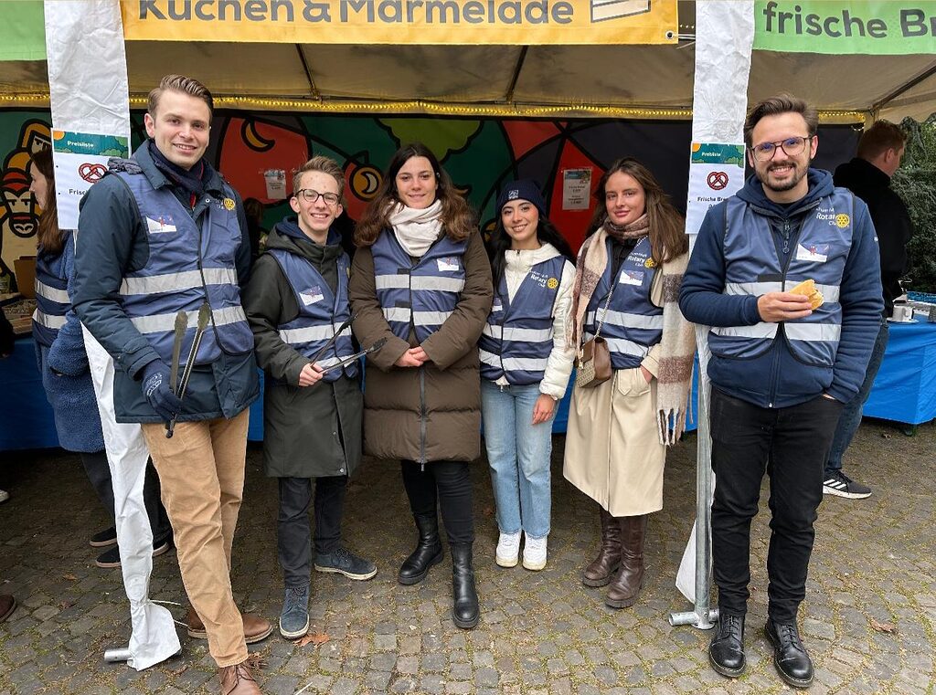 Sechs Rotaracter stehen in Winterkleidung und dunkelblauen Warnwesten vor einem Stand des St.-Martins-Marktes und gucken in die Kamera.