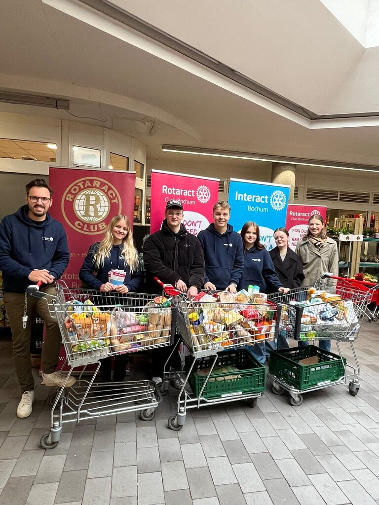 Rotaracter stehen gemeinsam mit Interactern vor drei prall gefüllten Einkaufswägen mit Lebensmitteln. In Hintergrund stehen Roll-ups mit den Logos von Rotaract und Interact. Teilweise halten sie Flyer in der Hand.